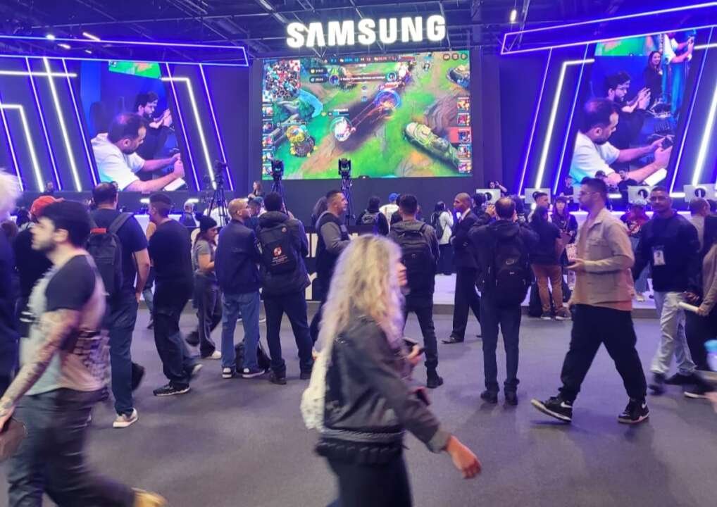 Aglomerado de pessoas transitando em frente ao estande da Samsung, onde ao fundo possui um telão que está sendo transmitido um campeonato local de League of Legends.